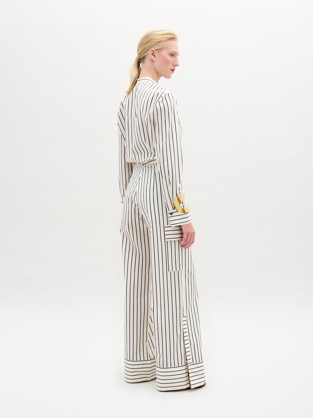 A person stands against a white background wearing a white long-sleeved blouse with vertical black stripes and lace detail, paired with the Falak Pant—relaxed fit, wide-leg trousers crafted from premium Italian fabric.