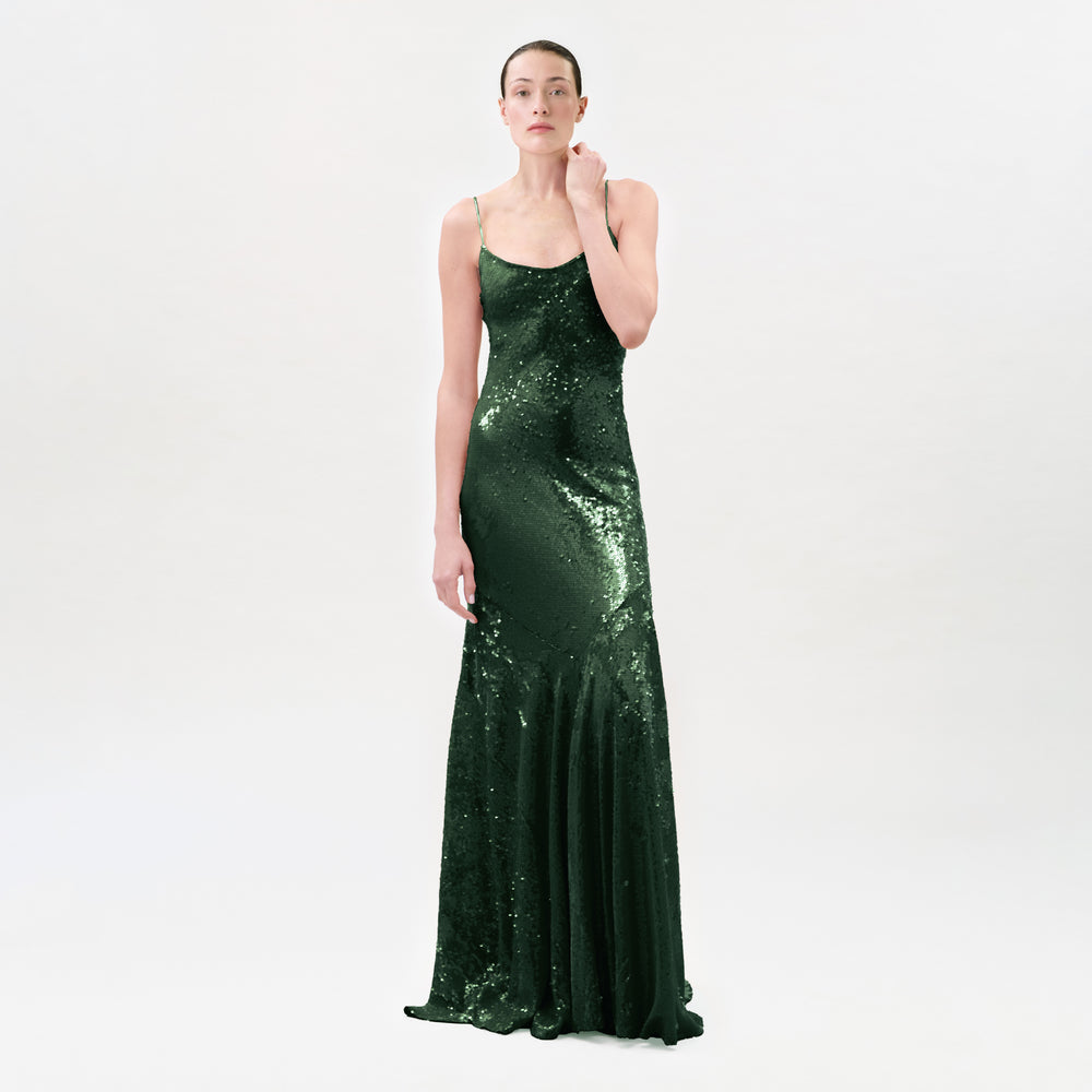 A woman modeling a floor-length, dark green sequined gown with thin spaghetti straps and a fitted silhouette, photographed against a seamless light gray studio background.