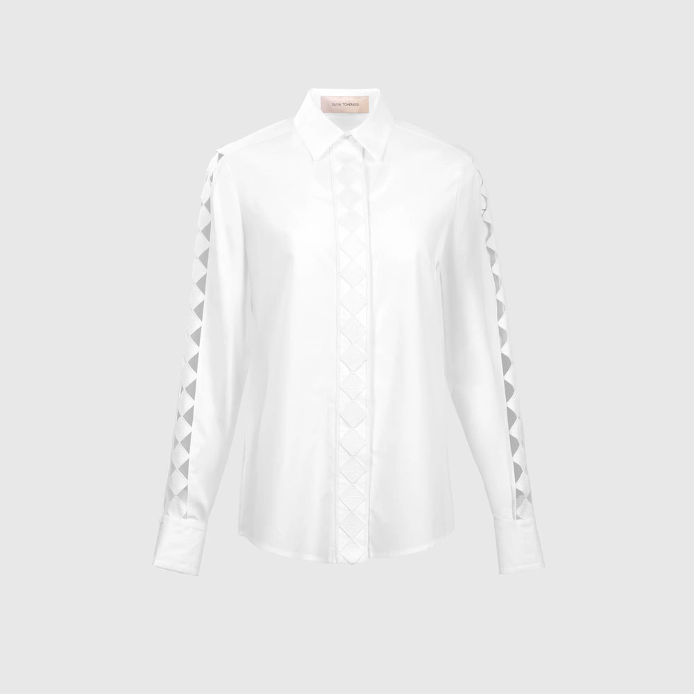 The Idris Blouse is a white long-sleeve top with a pointed collar, made from quality Portuguese fabric. It showcases a geometric triangular pattern on the sleeves and front placket, set against a neutral gray background.