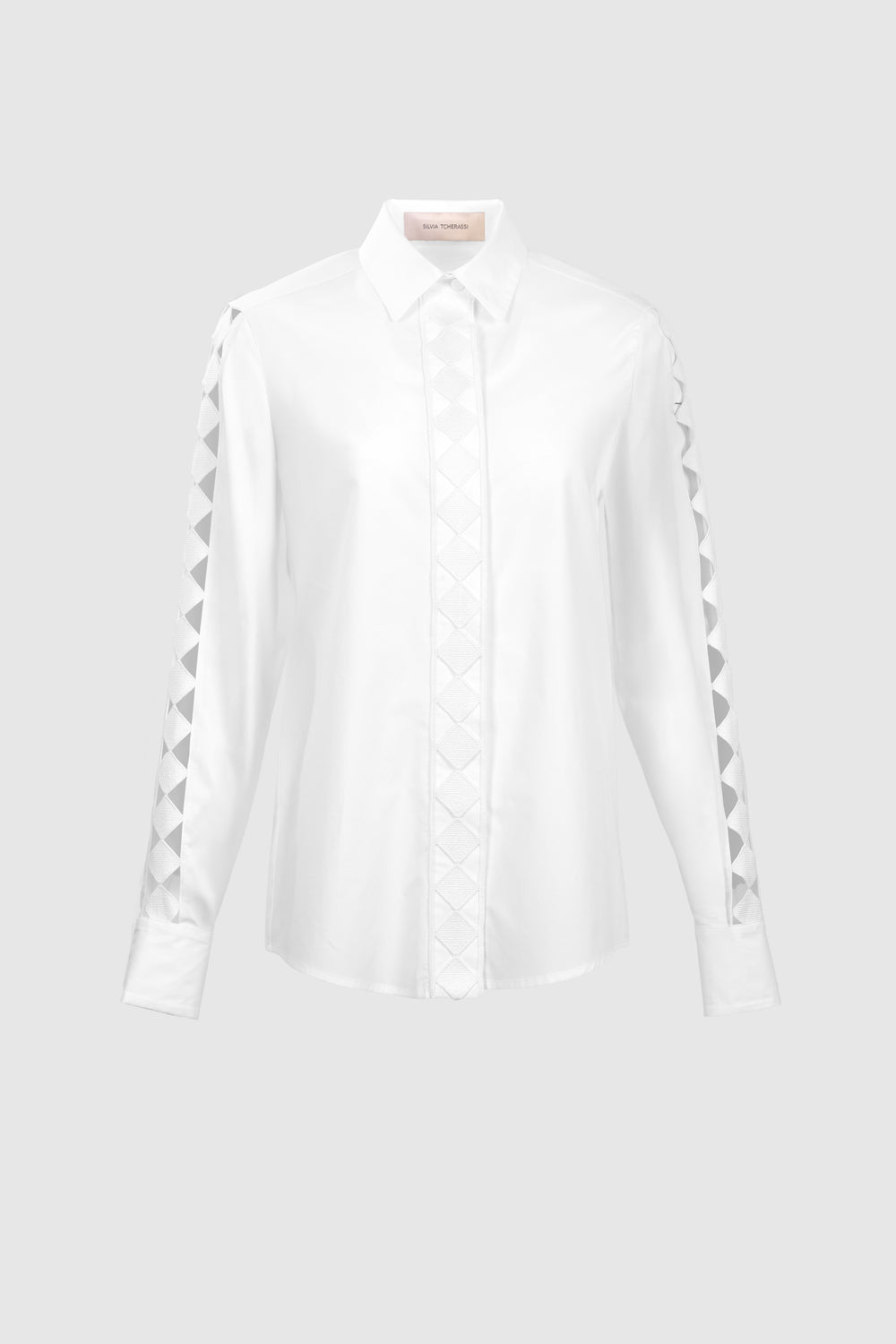 The Idris Blouse is a white, long-sleeve top made from 100% Portuguese cotton with a pointed collar and geometric triangle patterns on the sleeves and front placket, shown against a plain light gray background.