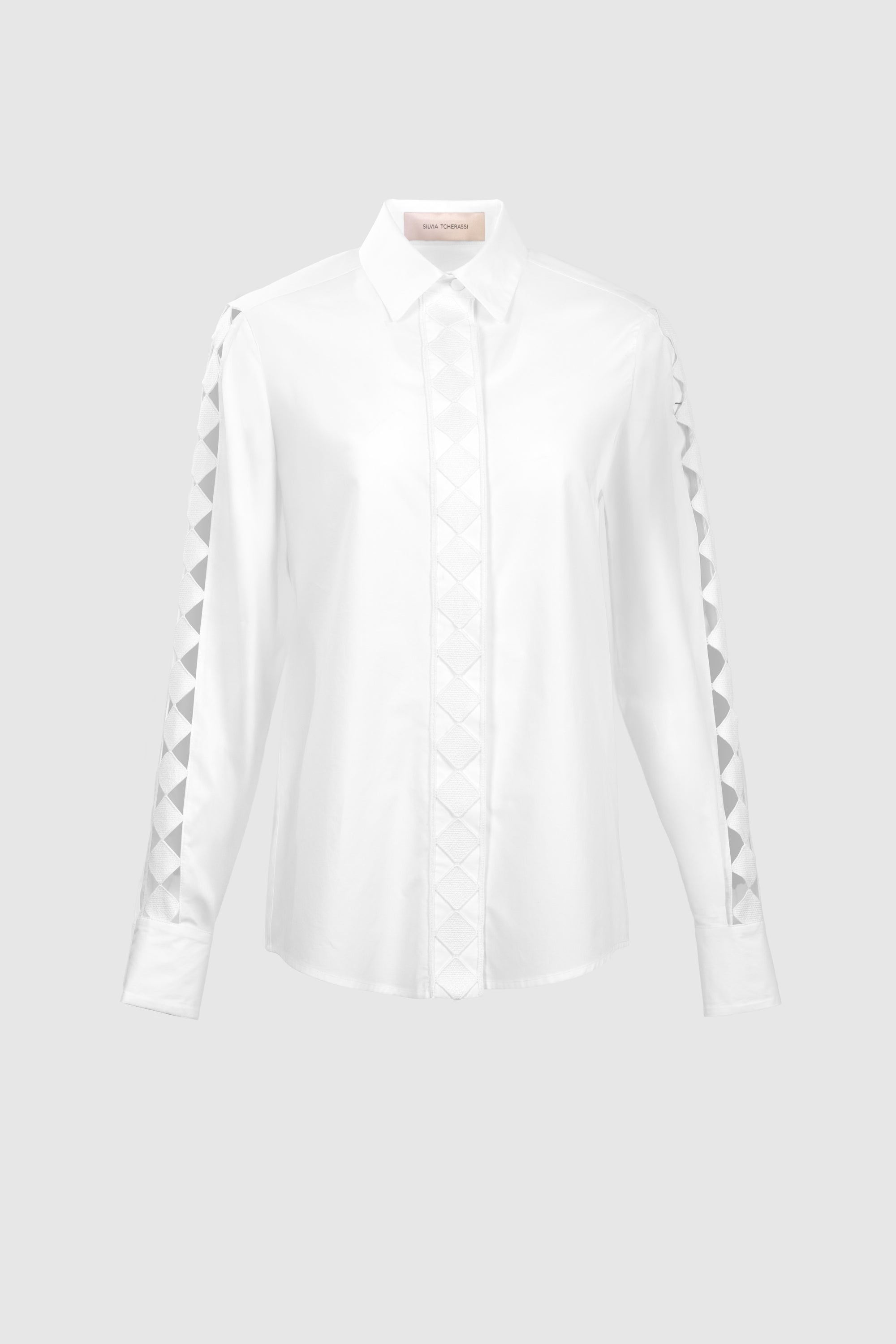 The Idris Blouse is a white, long-sleeve top made from 100% Portuguese cotton with a pointed collar and geometric triangle patterns on the sleeves and front placket, shown against a plain light gray background.