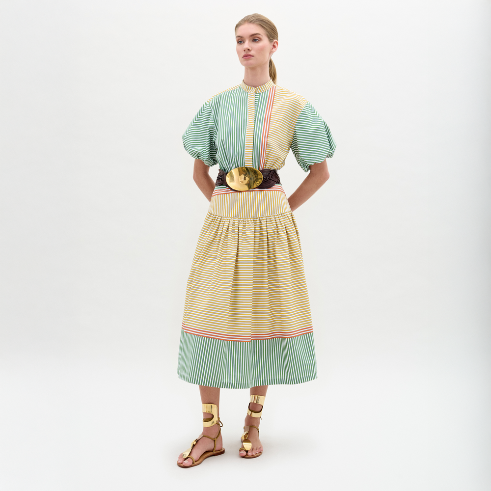 A woman stands against a plain background wearing the Jil Dress, a green and yellow striped midi with puffed sleeves, an A-line skirt, wide belt with large buckle, and gold sandals.