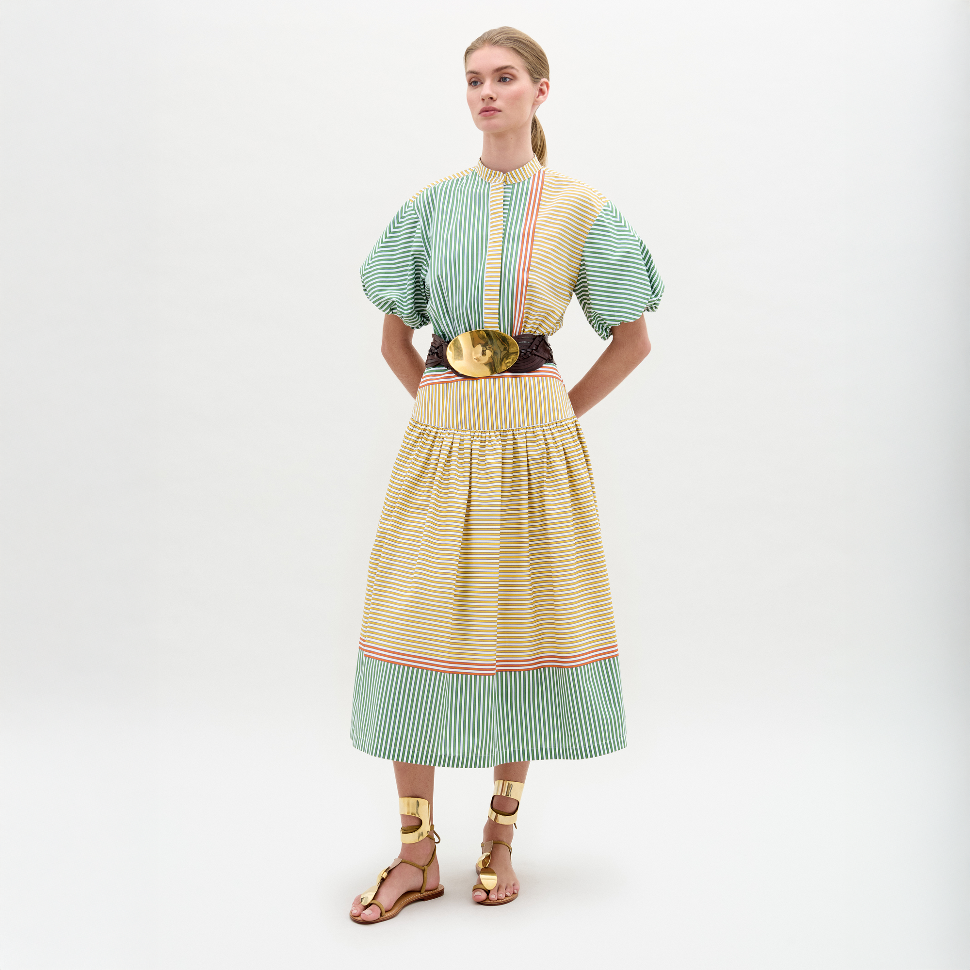 A woman stands against a plain background wearing the Jil Dress, a green and yellow striped midi with puffed sleeves, an A-line skirt, wide belt with large buckle, and gold sandals.