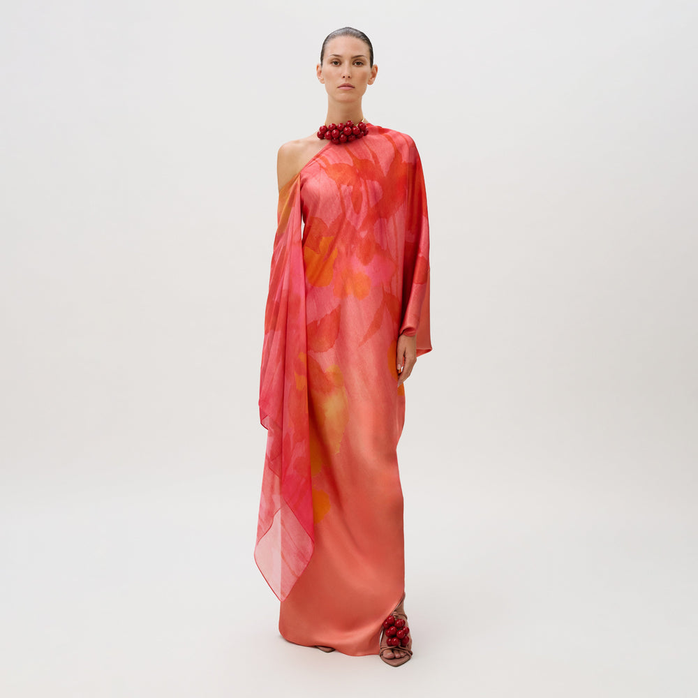 A model wears the Jade Dress, a floor-length, one-shoulder silk chiffon gown in coral and pink with a draped side panel, styled with a red beaded collar and matching beaded sandals.