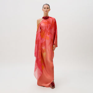 A model wears the Jade Dress, a floor-length, one-shoulder silk chiffon gown in coral and pink with a draped side panel, styled with a red beaded collar and matching beaded sandals.