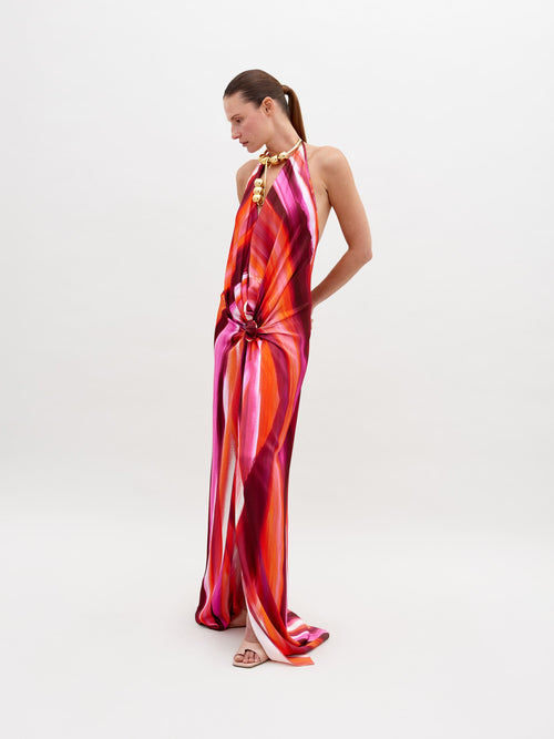 A woman stands against a plain background, wearing a colorful, flowing halter-neck Jazmin Dress Magenta Abstract Waves with a prominent knot detail at the waist. Available for pre-order now, the dress will ship by November 15th, 2024. She has her hands behind her back and is looking down.