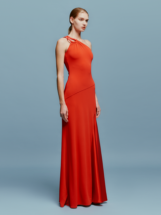 A woman in a Kanya Dress Rouge, featuring a red, one-shoulder design and floor-length cut, stands against a plain blue background. Her hair is pulled back as she calmly stands with her arms by her sides, appearing to wait for an important date such as August 19th or September 2nd.