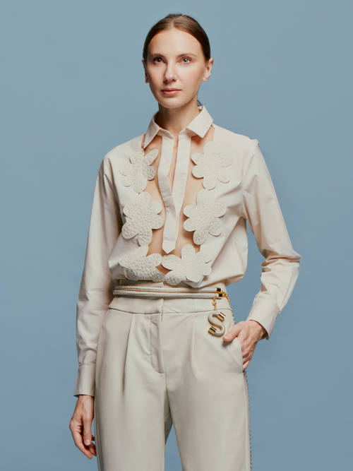 A person stands against a blue background, wearing the Karole Blouse Cream adorned with large floral decorations and holding a small accessory attached to their pants. Please note that this item’s shipping dates are between August 19th and September 2nd.