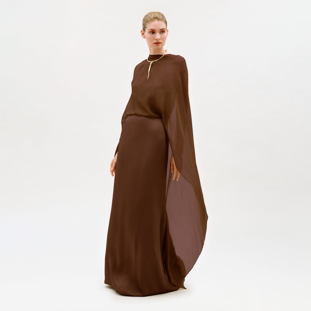 A woman wears the Kalina Dress, a long, flowing brown maxi gown featuring a cape detail and column silhouette, as she stands against a plain white background and gazes slightly to the side.