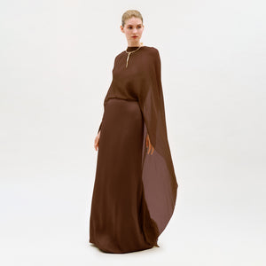 A woman wears the Kalina Dress, a long, flowing brown maxi gown featuring a cape detail and column silhouette, as she stands against a plain white background and gazes slightly to the side.