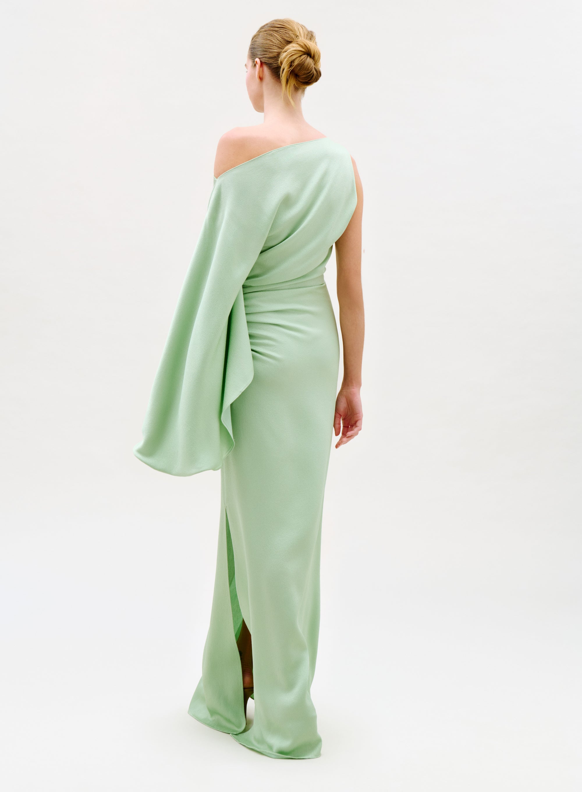 A woman faces away in the Kesia Dress, a pale green one-shoulder gown with a long draped sleeve, her hair styled in a neat bun against a plain white background.