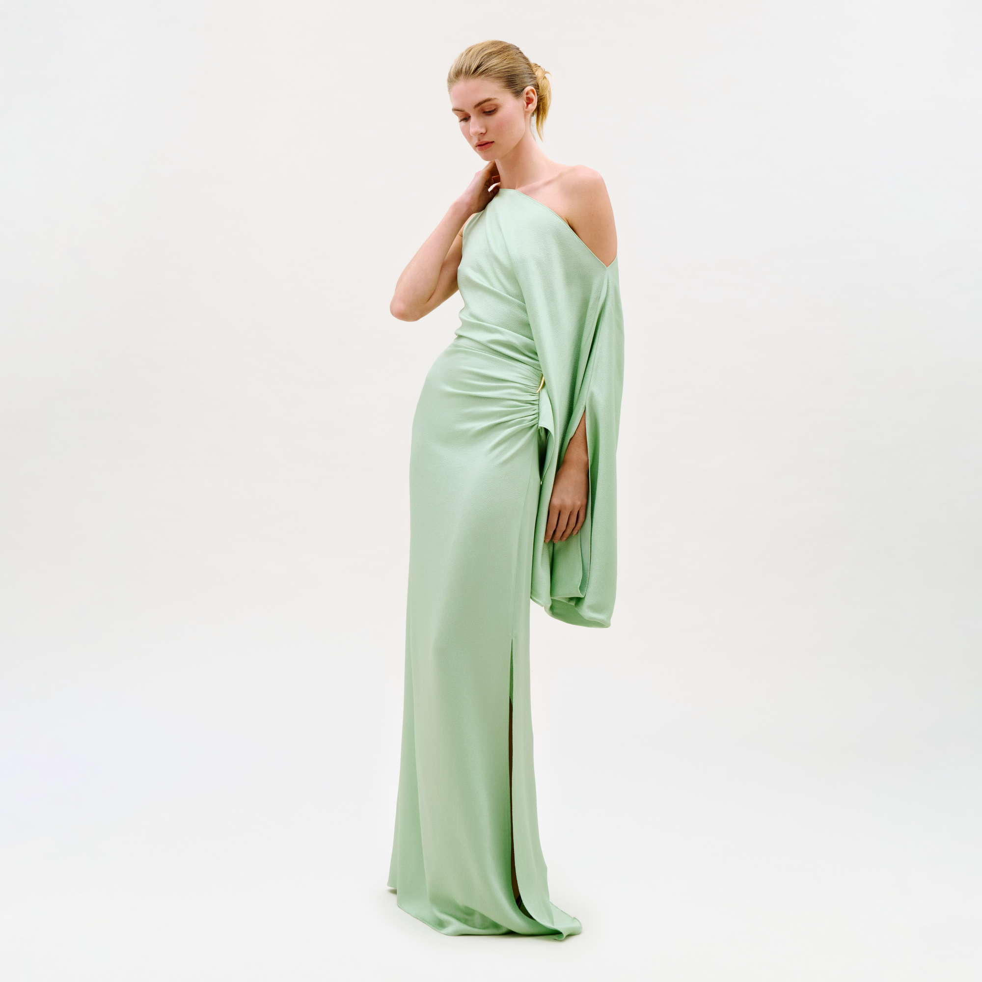 A woman wears the Kesia Dress, a light green off-the-shoulder floor-length gown, against a plain light background, looking down with one hand on her neck and the other at her side.