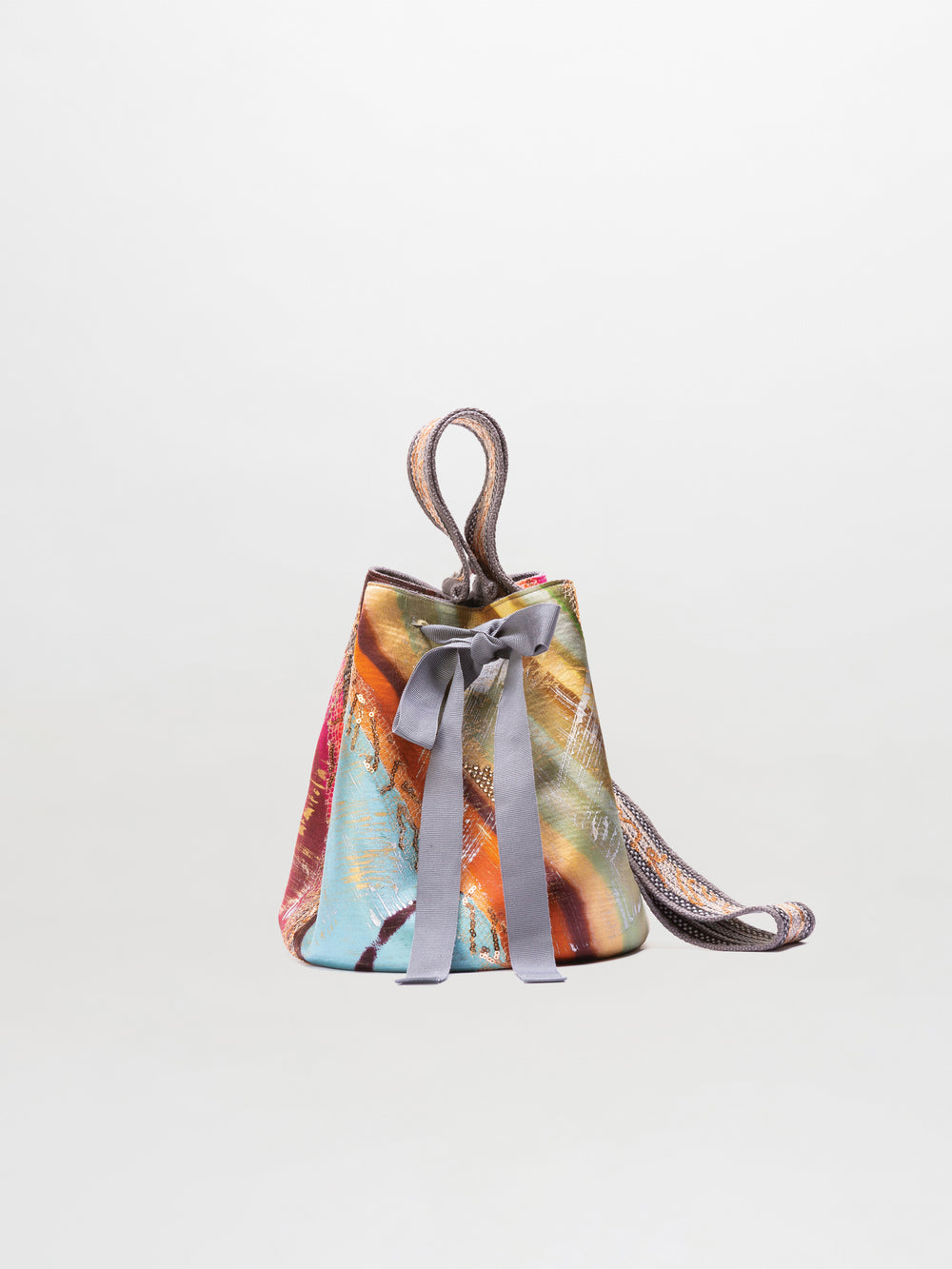 Introducing the Mochila Laaput Small: a vibrant, patterned small bucket bag crafted in Colombia, adorned with a decorative grey ribbon bow at the front and finished with a multicolored shoulder strap, shown against a plain white background.