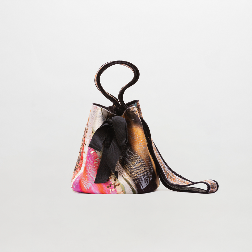 The Mochila Laaput Big is a small bucket-style Wayuu handbag featuring a multicolored pattern, top handle, black ribbon bow, and hand-woven strap, shown against a plain white background.