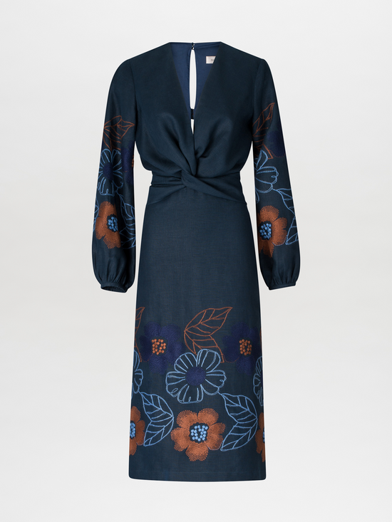 A person stands against a blue background wearing the Laoise Dress in Navy Ochre Flower Embroidery, paired with beige high-heeled sandals, perfect for an August 19th or September 2nd outing.