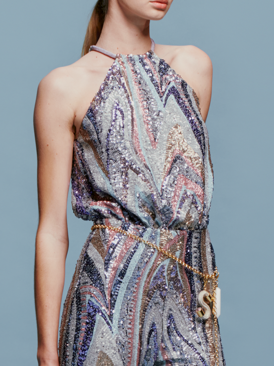 Against a plain blue background, a person stands poised in the Lauretta Jumpsuit Sequin Waves, featuring a sleeveless, high-neck design with a colorful, swirling pattern. The outfit showcases wide-leg pants and comes with a belt—perfectly capturing the style for upcoming events between August 19th and September 2nd, 2024.