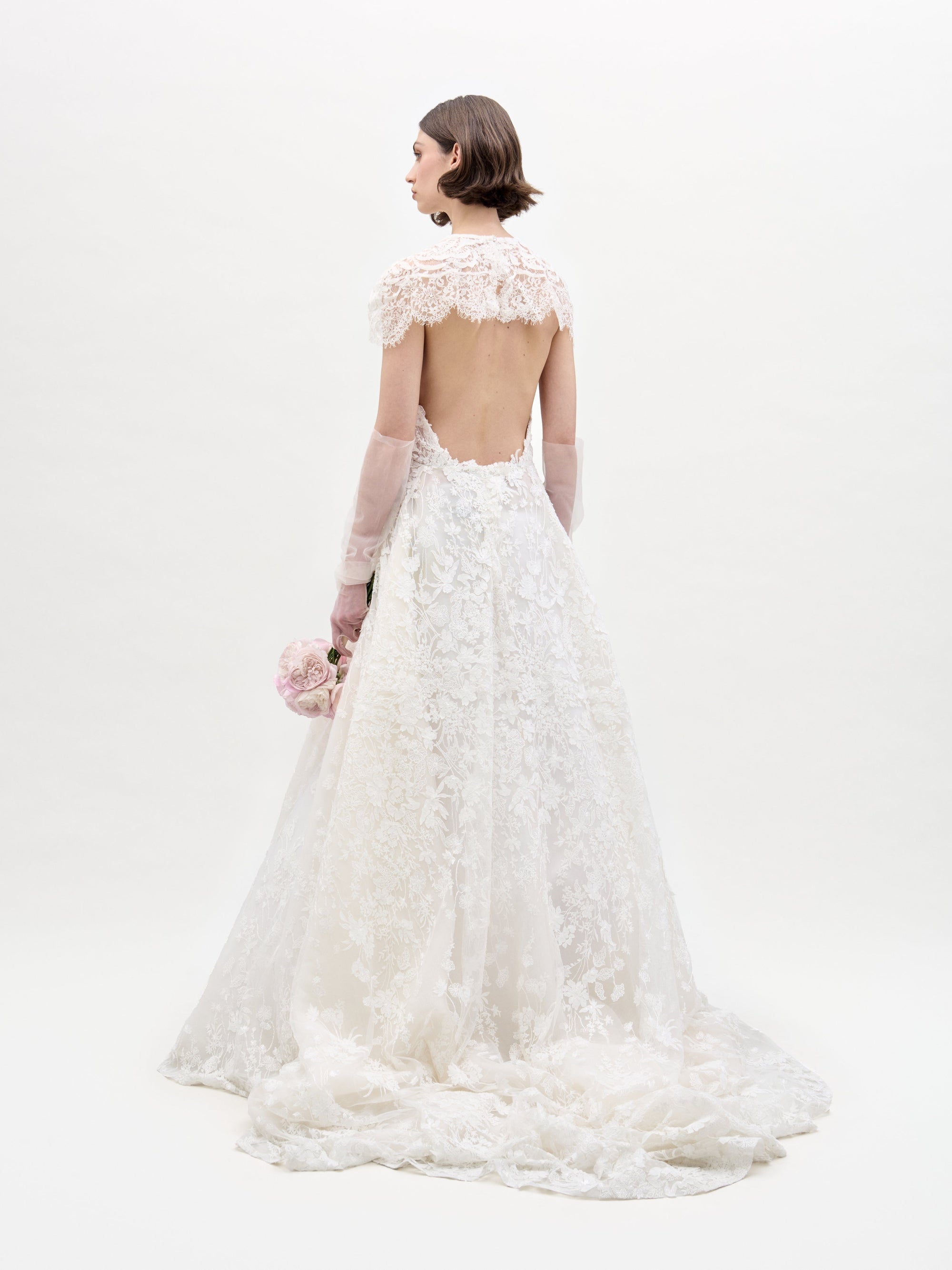 A woman stands facing away on a plain white background, holding pink flowers and wearing the Leia Dress—a white, lacy, backless wedding maxi dress with elegant lace details and a long train.