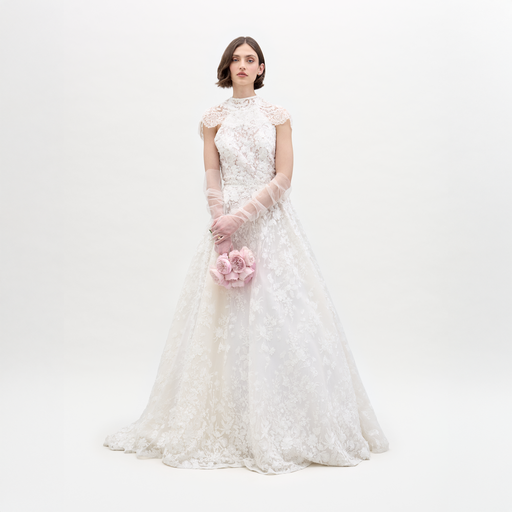A woman wears the Leia Dress, a white high-neck lace wedding gown with delicate detailing and sheer gloves, holding a bouquet of pale pink roses against a plain white background.