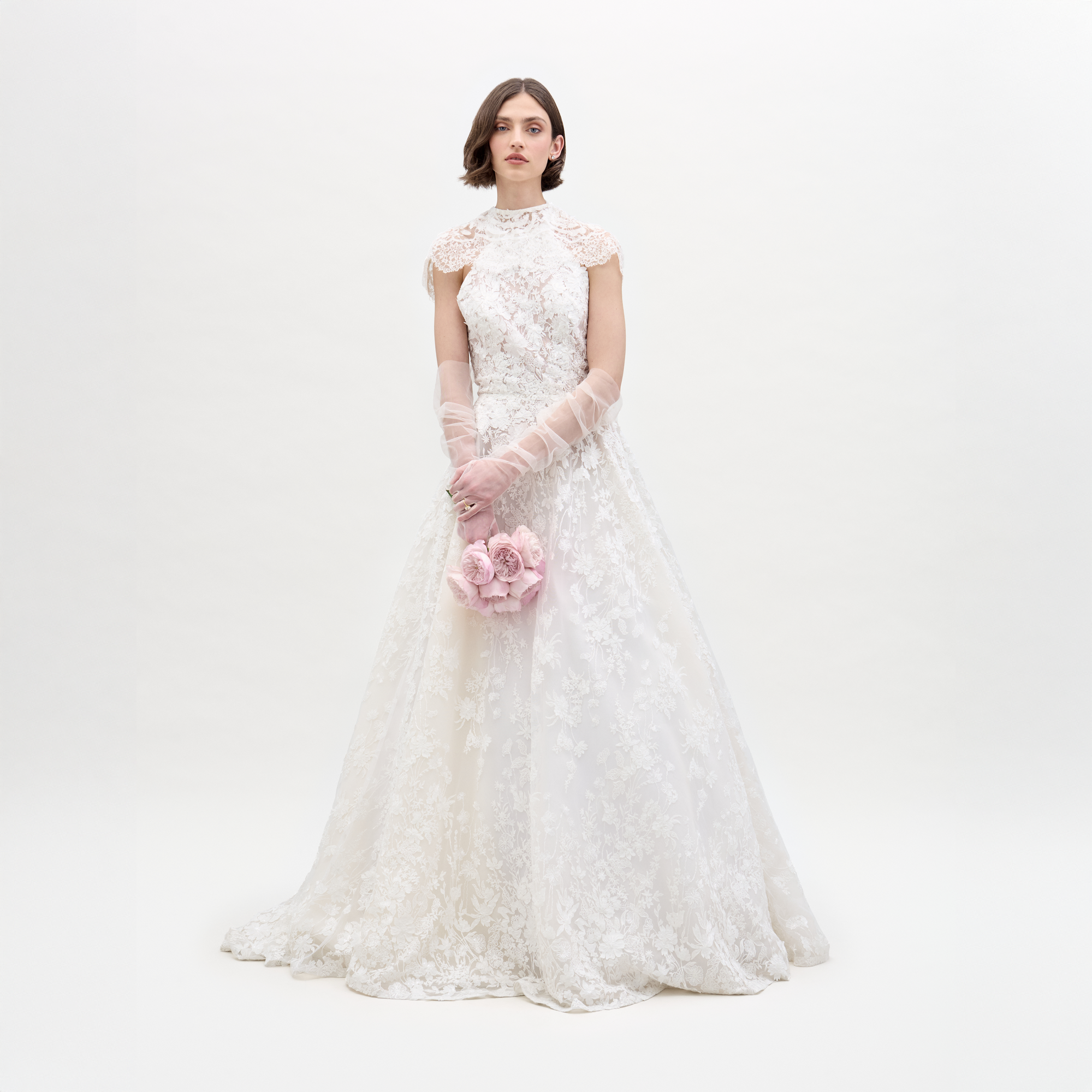 A woman wears the Leia Dress, a white high-neck lace wedding gown with delicate detailing and sheer gloves, holding a bouquet of pale pink roses against a plain white background.