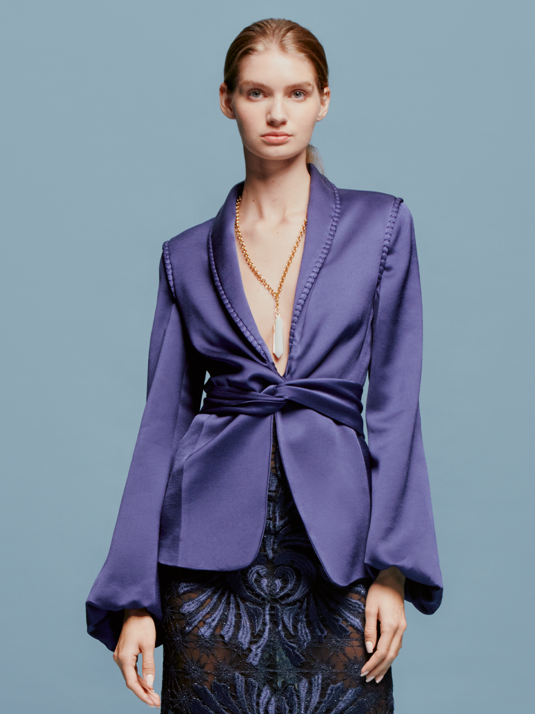 A person in a chic Leo Jacket Navy and a patterned skirt stands against a light blue background, embodying the stylish essence of September 2nd. They are wearing a long necklace with a white pendant.