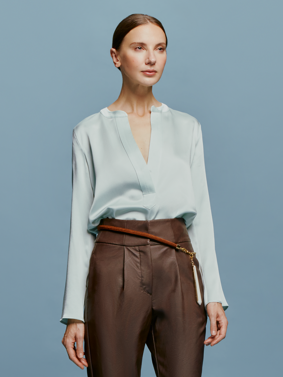 A woman stands against a plain blue background, wearing the Lilly Blouse in Turquoise and brown high-waisted pants, with a relaxed and composed expression, as if ready for September 2nd, 2024.