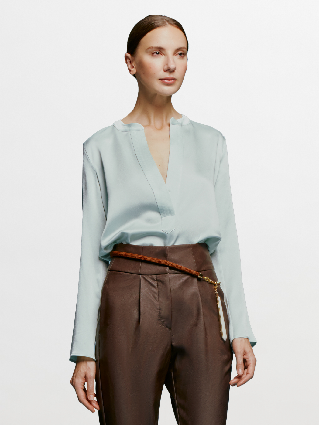 A woman stands against a plain blue background, wearing the Lilly Blouse in Turquoise and brown high-waisted pants, with a relaxed and composed expression, as if ready for September 2nd, 2024.