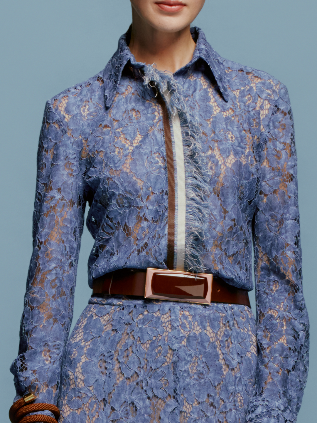 A woman stands against a plain blue background, wearing the Liv Blouse in Blue. She pairs it with a stylish brown bracelet, perfectly capturing the essence of September 2nd, 2024.