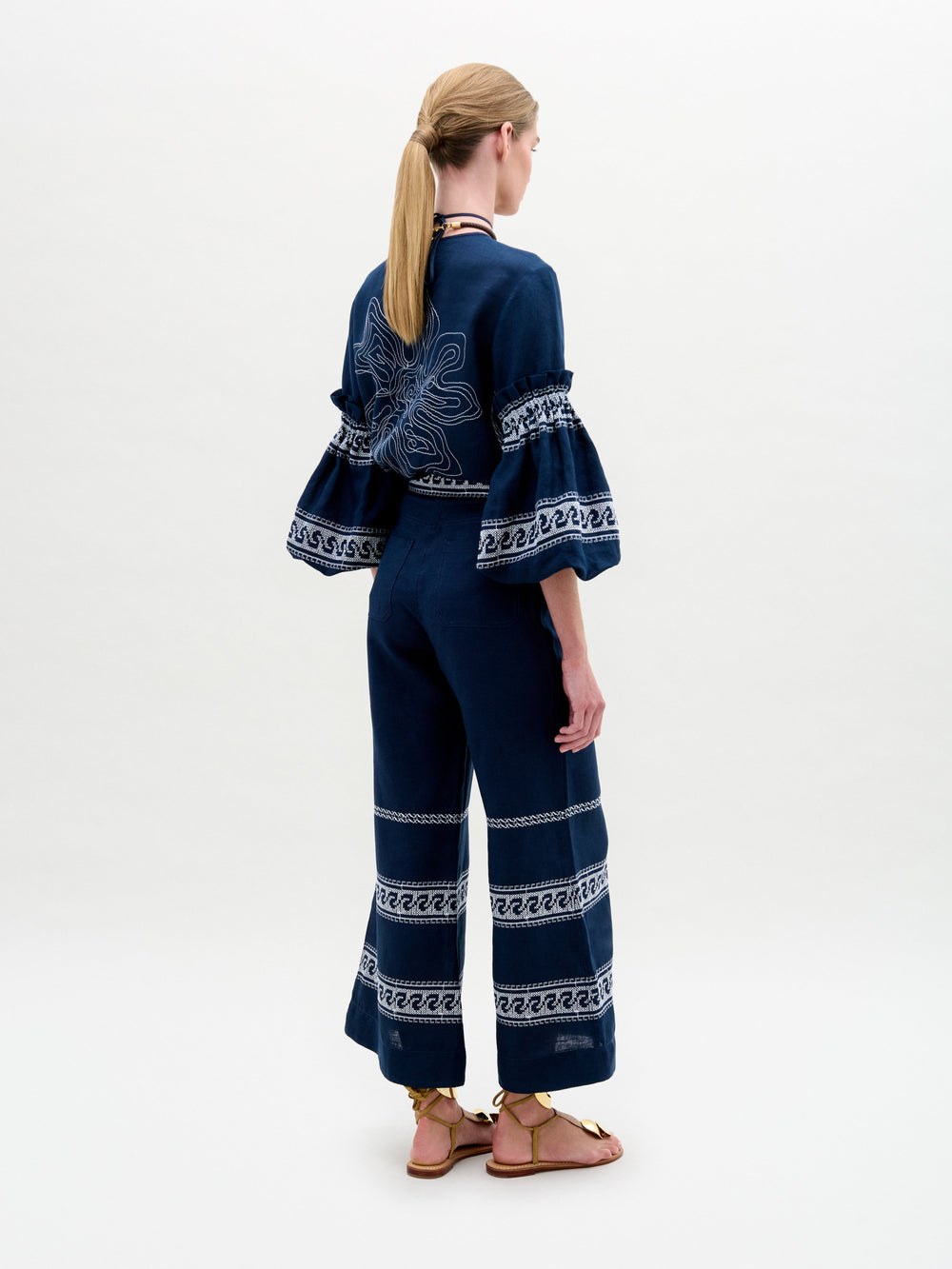 A woman stands against a plain background, wearing a navy blue embroidered blouse with ruffled sleeves and the Zina Pant—matching embroidered linen wide-leg pants—paired with gold sandals.