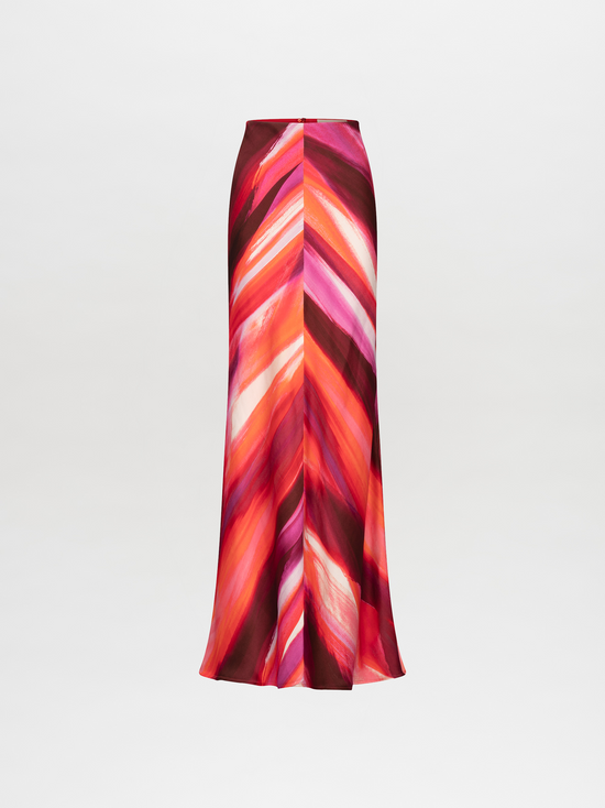 A person stands against a plain background, wearing a vibrant suit in red, pink, and orange streaks, paired with the Laurina Skirt Magenta Abstract Waves and bold gold necklace. This stunning ensemble will be available for pre-order soon and is set to ship by date 2024.