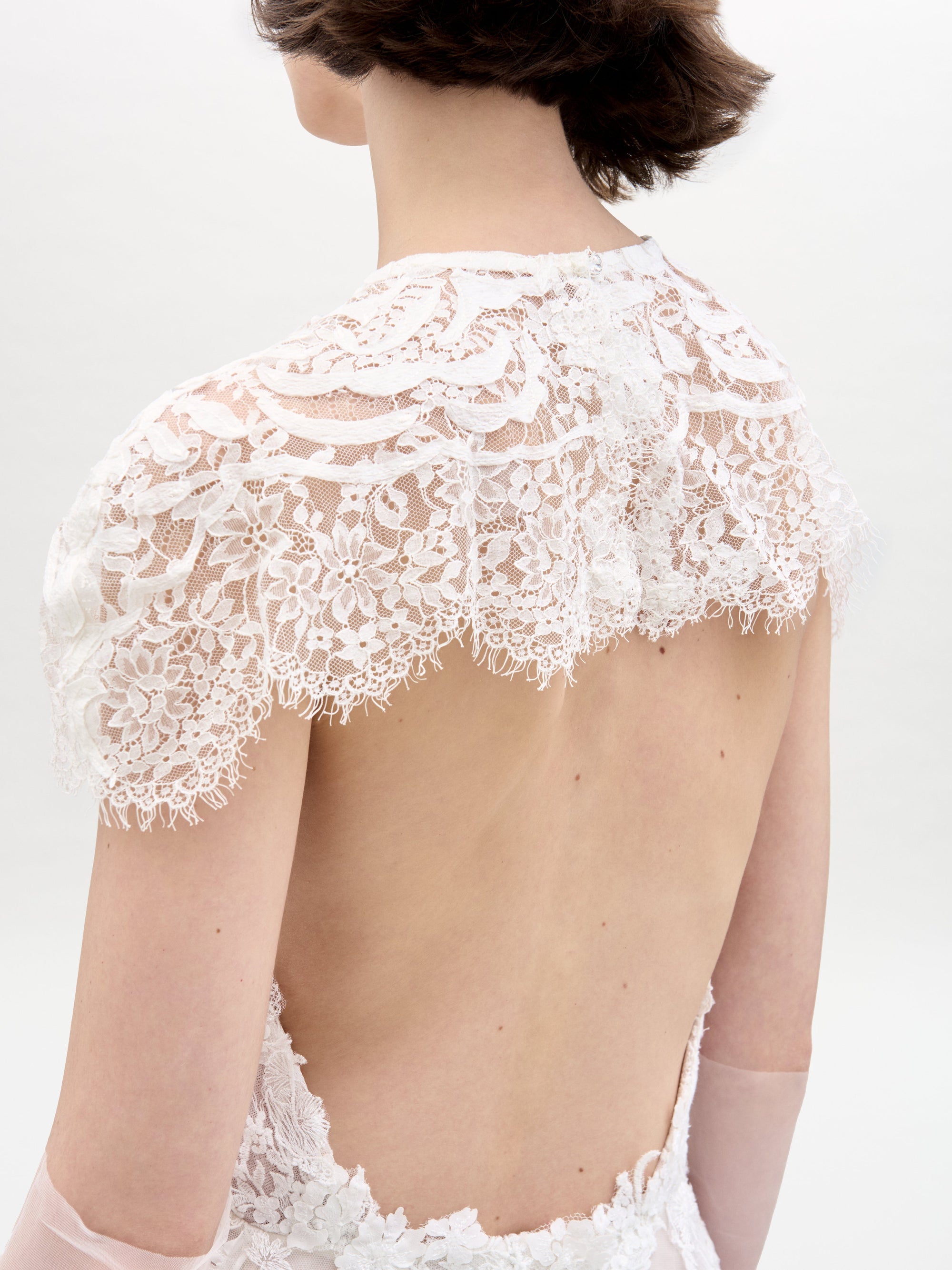 A woman with short brown hair models the Leia Dress, a white lace dress featuring elegant floral patterns, against a plain light background.