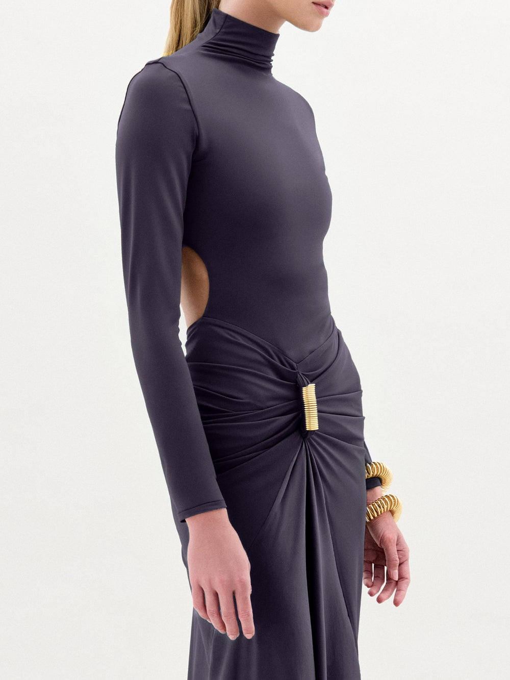 A person wearing the Lena Dress—a long-sleeved, high-neck, dark fitted dress with cut-out sides—paired with gold wrist accessories, stands against a plain light background.
