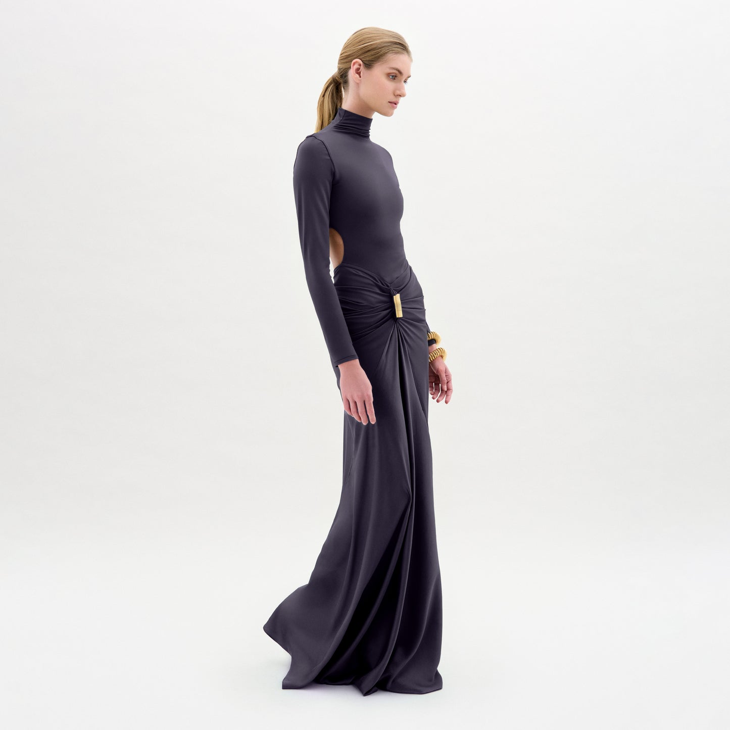 A woman stands in profile against a white background wearing the Lena Dress, a long, dark, form-fitting gown with long sleeves and side cutouts. Her hair is tied back and she accessorizes with gold bracelets.