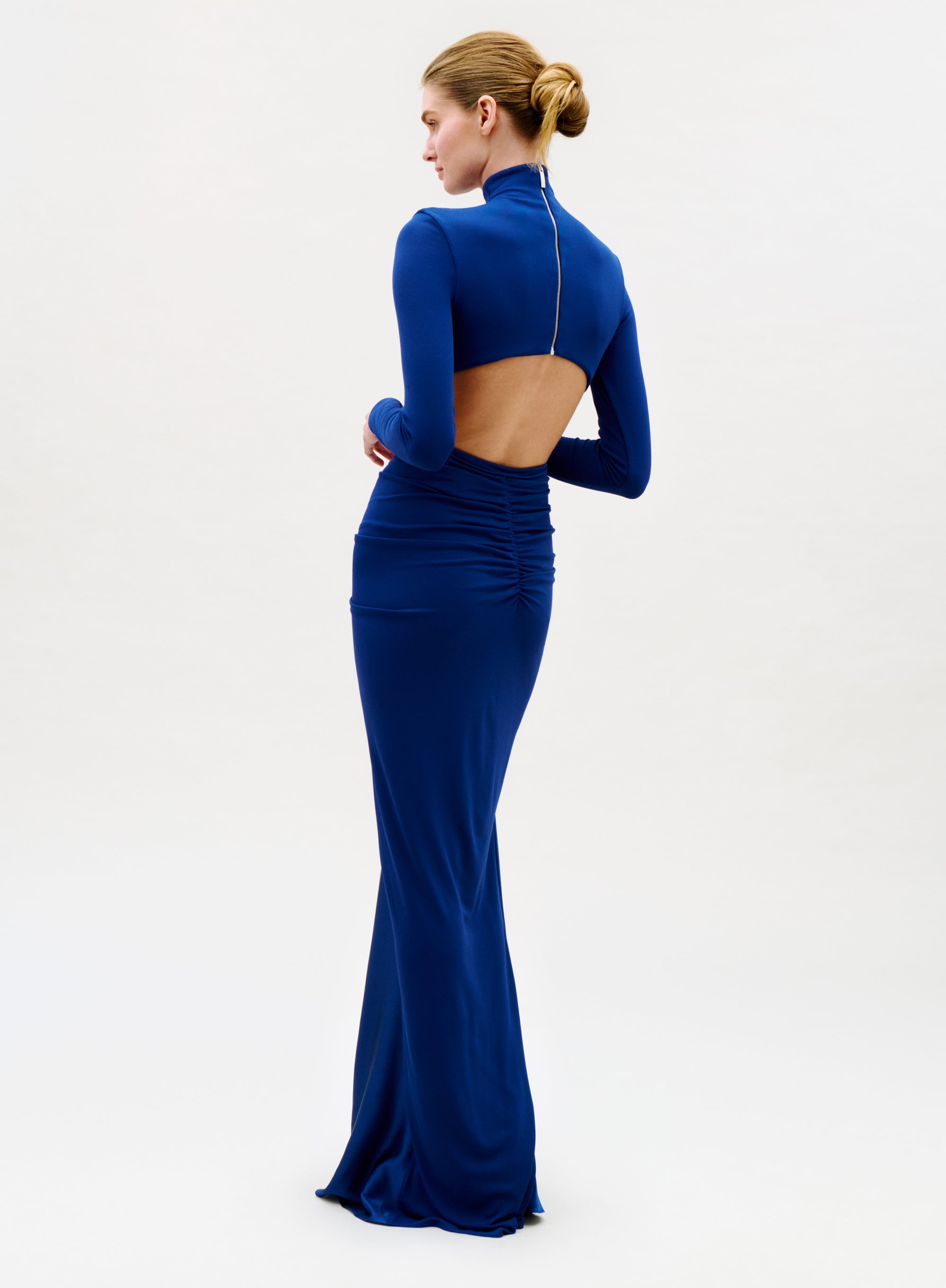 A woman faces away from the camera in the Lena Dress, a fitted, floor-length royal blue gown with long sleeves, an open back, and a visible zipper running from neck to lower back.