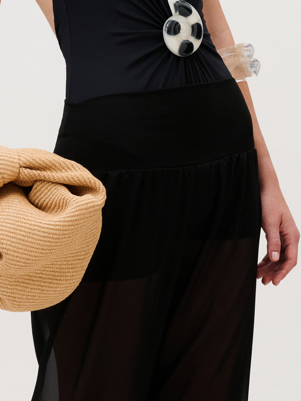 A person wearing a black sleeveless top with a large circular pendant and sheer chiffon Lene Pant, holding a beige woven clutch.