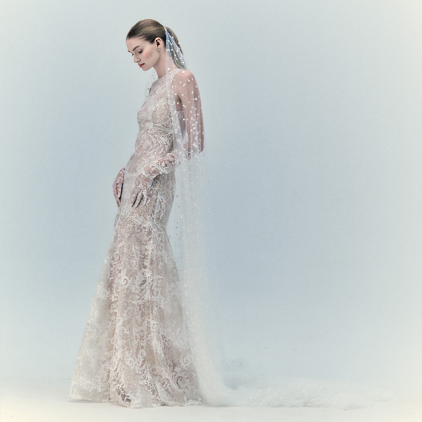A woman stands in profile wearing the Levi Dress, an elegant long lace wedding gown with sheer sleeves and embroidered florals, paired with a matching veil adorned with pearls, set against a soft, light background.