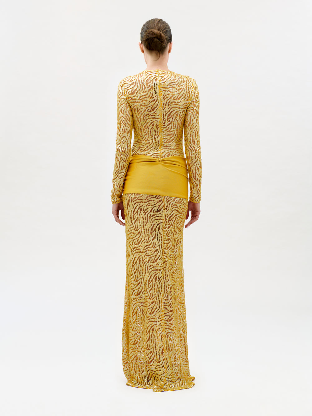 A person stands facing away, wearing the Lia Dress—a slim fit, long-sleeved, floor-length yellow gown crafted from luxurious Italian fabric featuring intricate sheer wavy patterns and a solid yellow panel around the hips. The background is plain and light-colored.