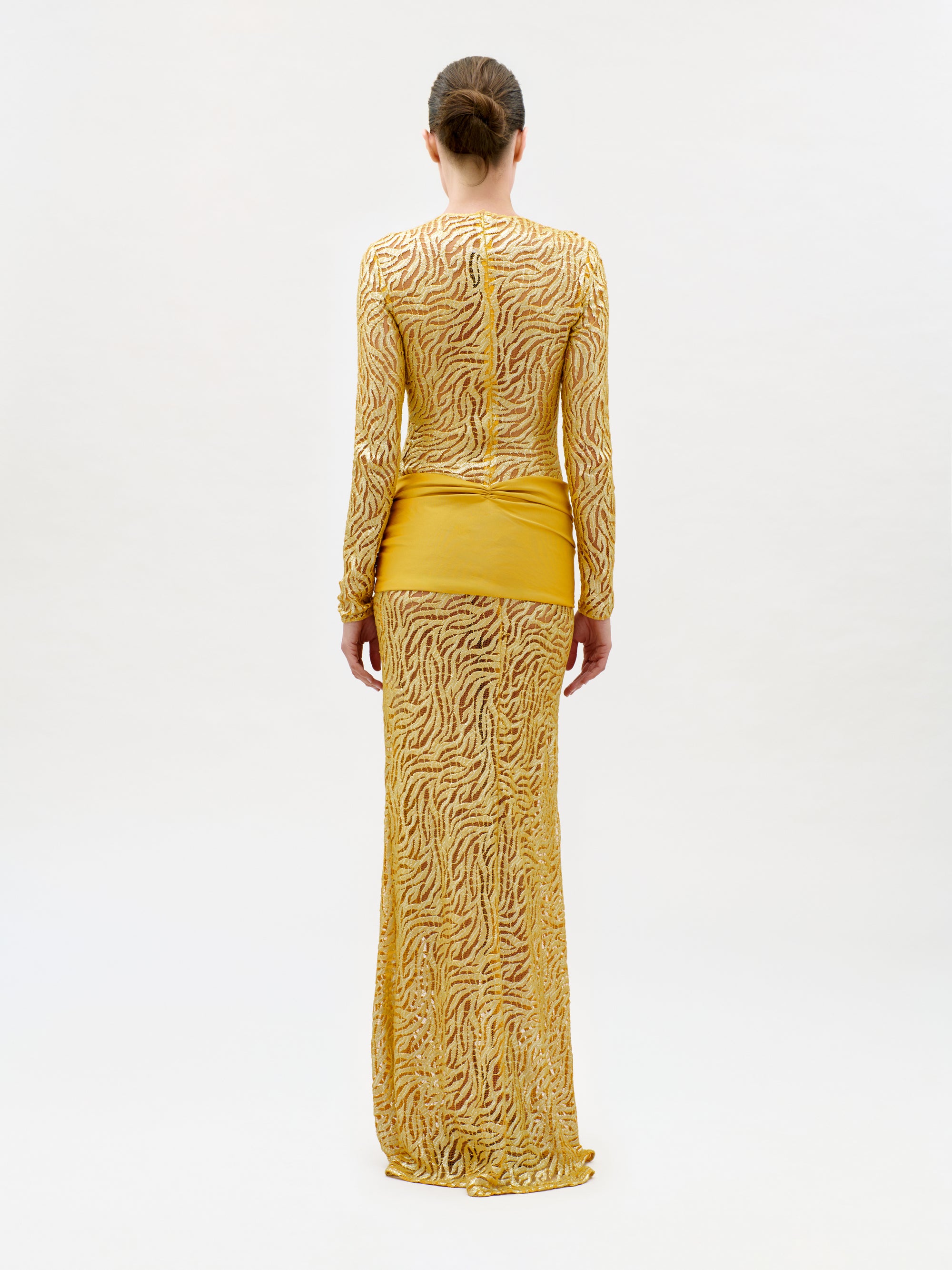 A person stands facing away, wearing the Lia Dress—a slim fit, long-sleeved, floor-length yellow gown crafted from luxurious Italian fabric featuring intricate sheer wavy patterns and a solid yellow panel around the hips. The background is plain and light-colored.