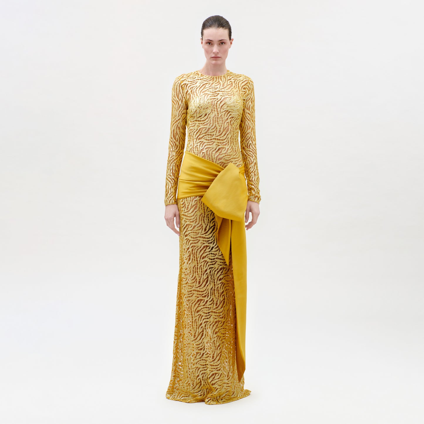 A woman wears the Lia Dress, a long-sleeved gold patterned gown made from luxurious Italian fabric, featuring a dramatic yellow draped sash at the waist, as she stands against a plain light background.