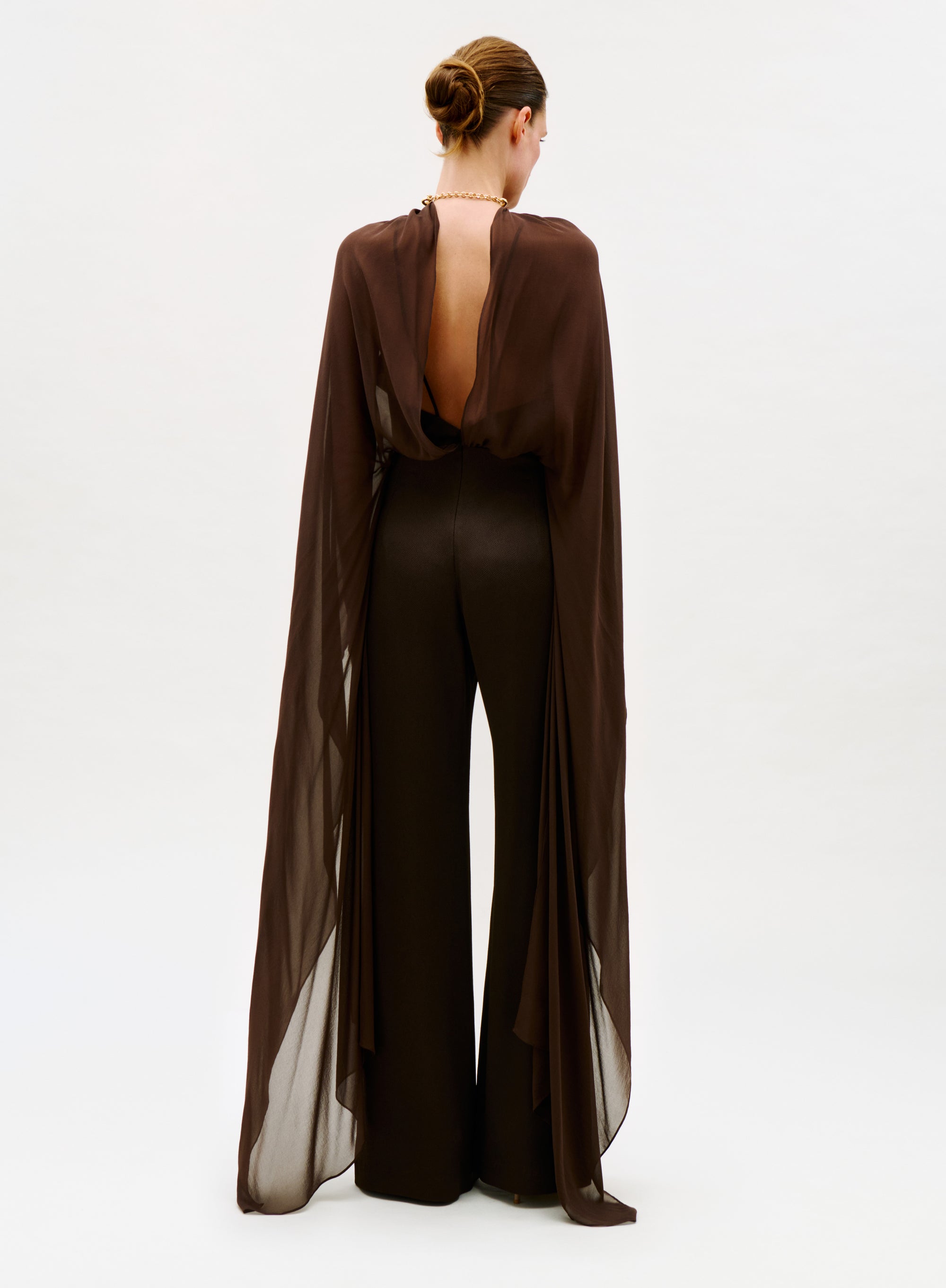 A person stands facing away, wearing the Ligia Jumpsuit—a sheer, flowing brown design with matching wide-leg pants and an open back—styled with a neat updo against a plain light background.