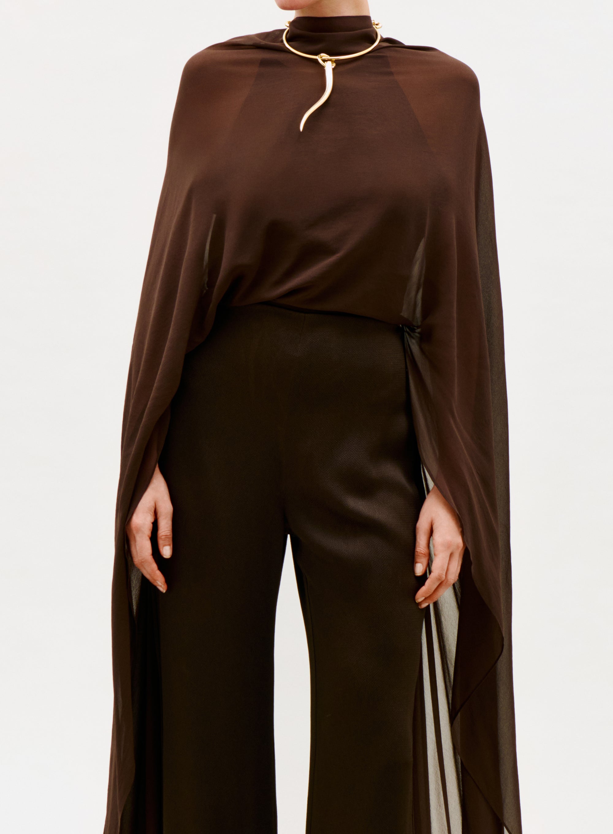 A person wears the Ligia Jumpsuit, a dark brown, semi-sheer piece with wide-leg pants, styled with a gold snake-shaped choker necklace. The background is plain white.