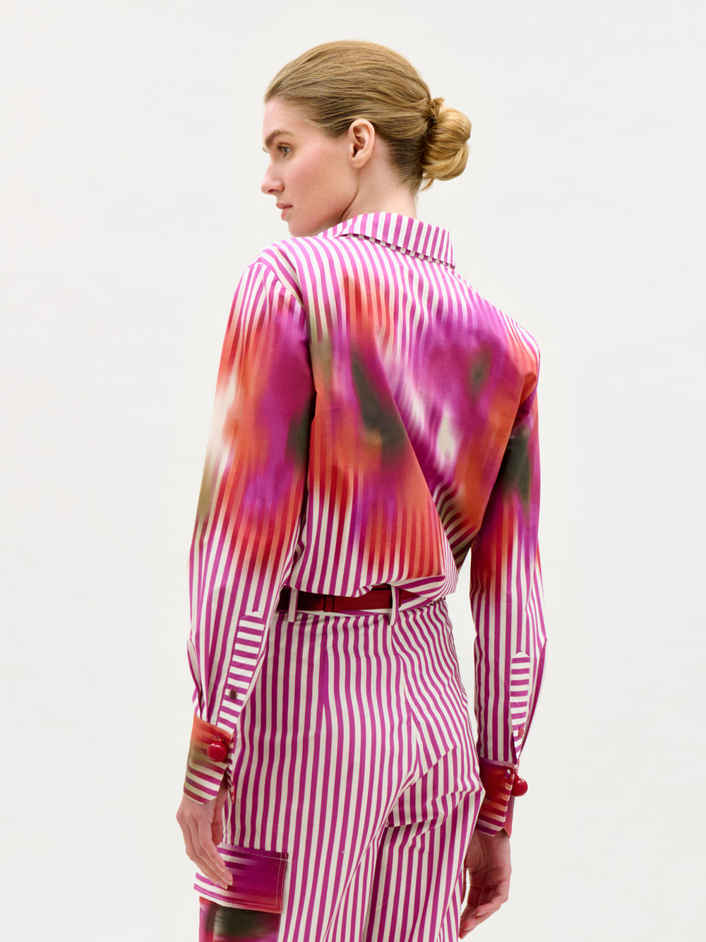 A woman with blonde hair in a low bun is seen from the back wearing the Linette Blouse—long-sleeved, made from Italian fabric, with bold pink and white stripes and colorful abstract patterns—against a plain white background.