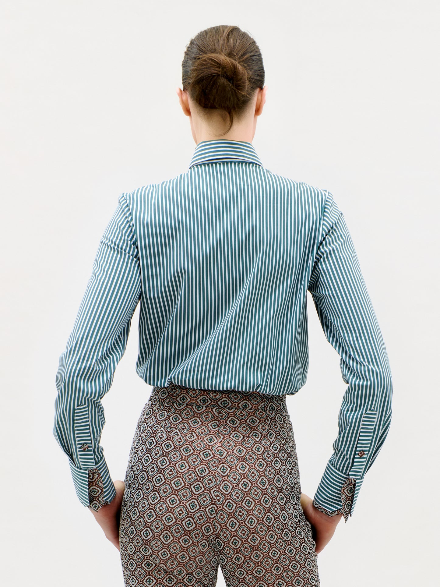 A person with brown hair in a bun faces away, wearing the Linnea Shirt—a blue and white vertical striped shirt—paired with patterned pants, against a plain white background.