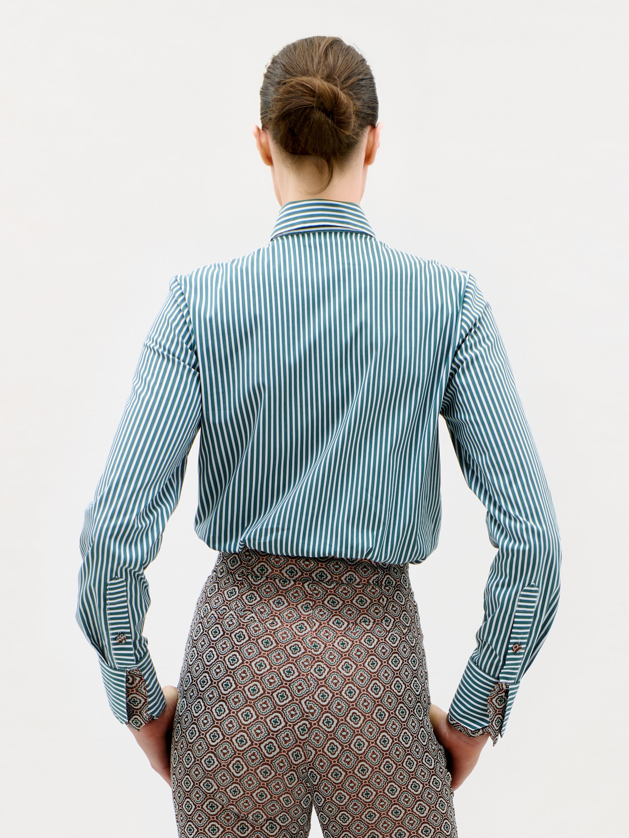 A person with brown hair in a bun faces away, wearing the Linnea Shirt—a blue and white vertical striped shirt—paired with patterned pants, against a plain white background.