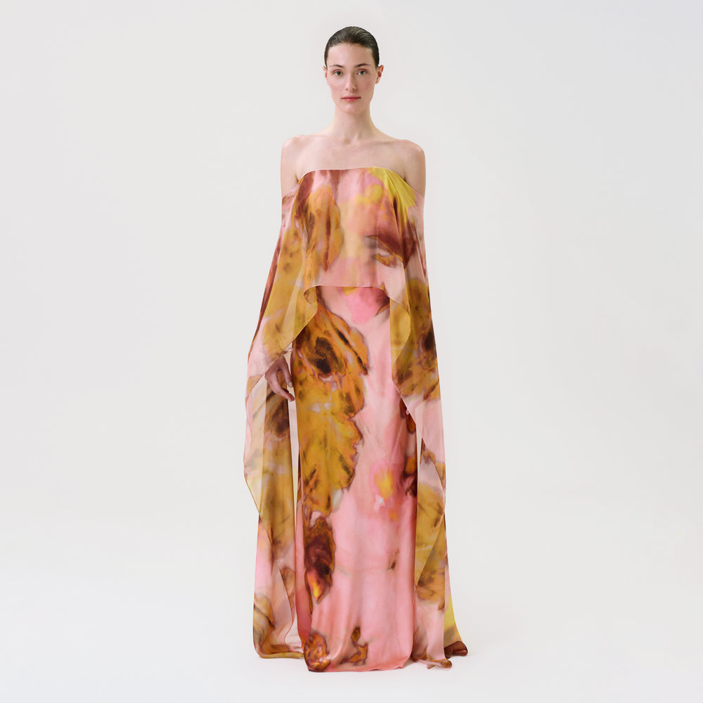 A model wearing the Lovane Dress, a strapless, floor-length gown with a loose, draped overlay featuring a pink, yellow, and brown abstract floral print.