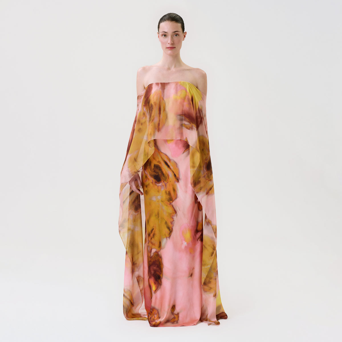 A model wearing the Lovane Dress, a strapless, floor-length gown with a loose, draped overlay featuring a pink, yellow, and brown abstract floral print.