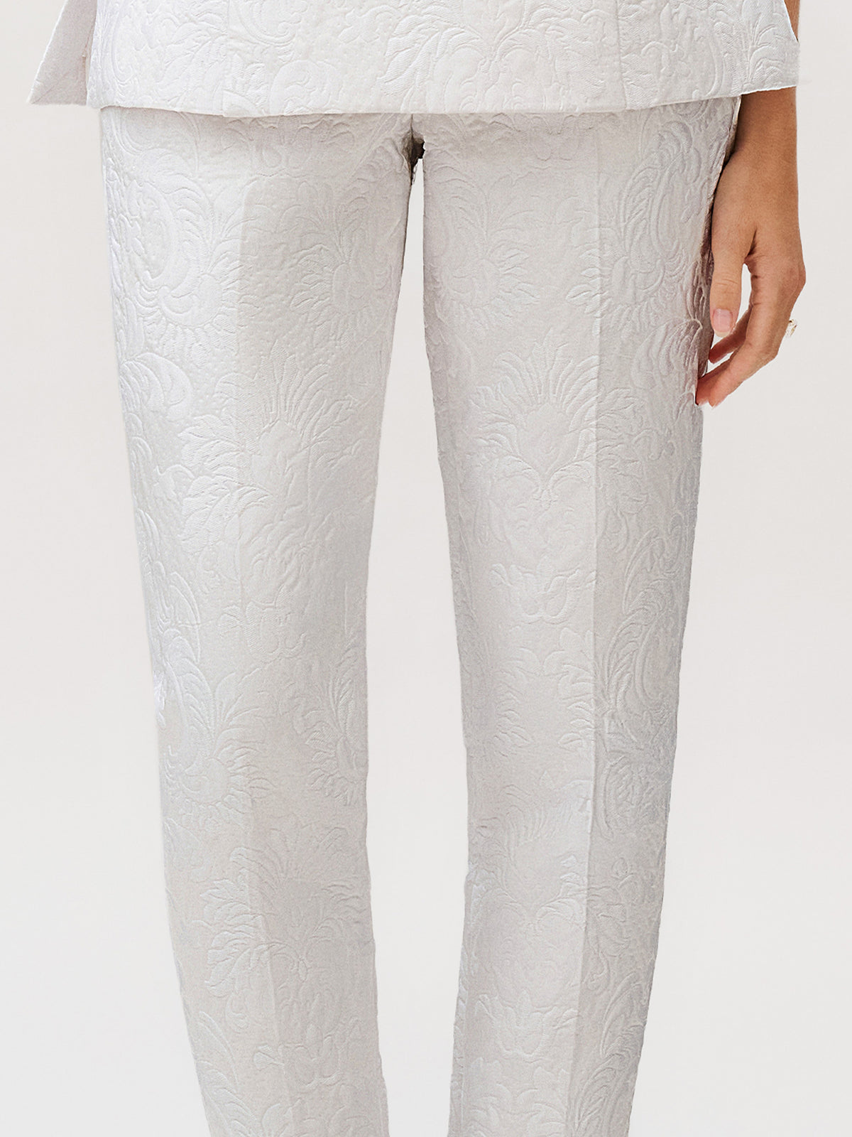A person wearing the Luca Pant in light gray silk cotton brocade with a textured floral pattern, paired with a matching jacket—an elegant ready-to-wear ensemble made in Italy, with one hand visible by their side.