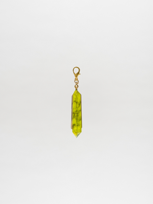 The Mariela Charm Green, by Tcherassi, is elegantly displayed against a plain white background, showcasing a green gemstone pendant with a gold clasp and an enchanting crystal charm.