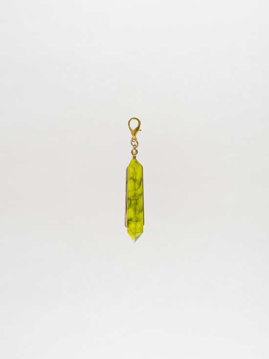 The Mariela Charm Green, by Tcherassi, is elegantly displayed against a plain white background, showcasing a green gemstone pendant with a gold clasp and an enchanting crystal charm.