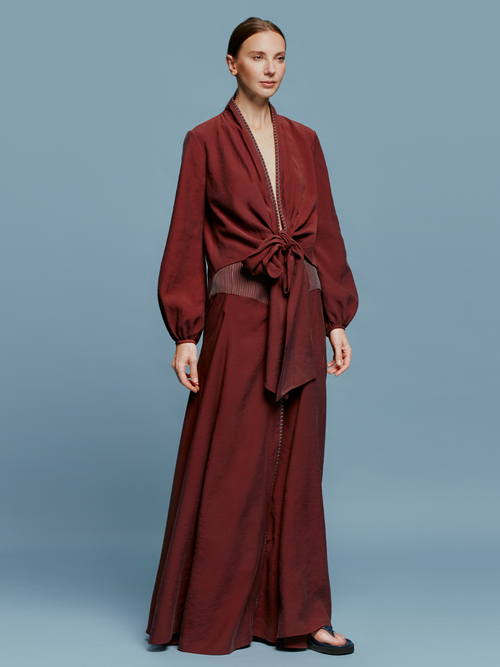 A person in a Modena Skirt Burgundy outfit, consisting of a long-sleeved top with a front tie and a matching long skirt, stands against a plain blue background, evoking memories of summer days on the ship.