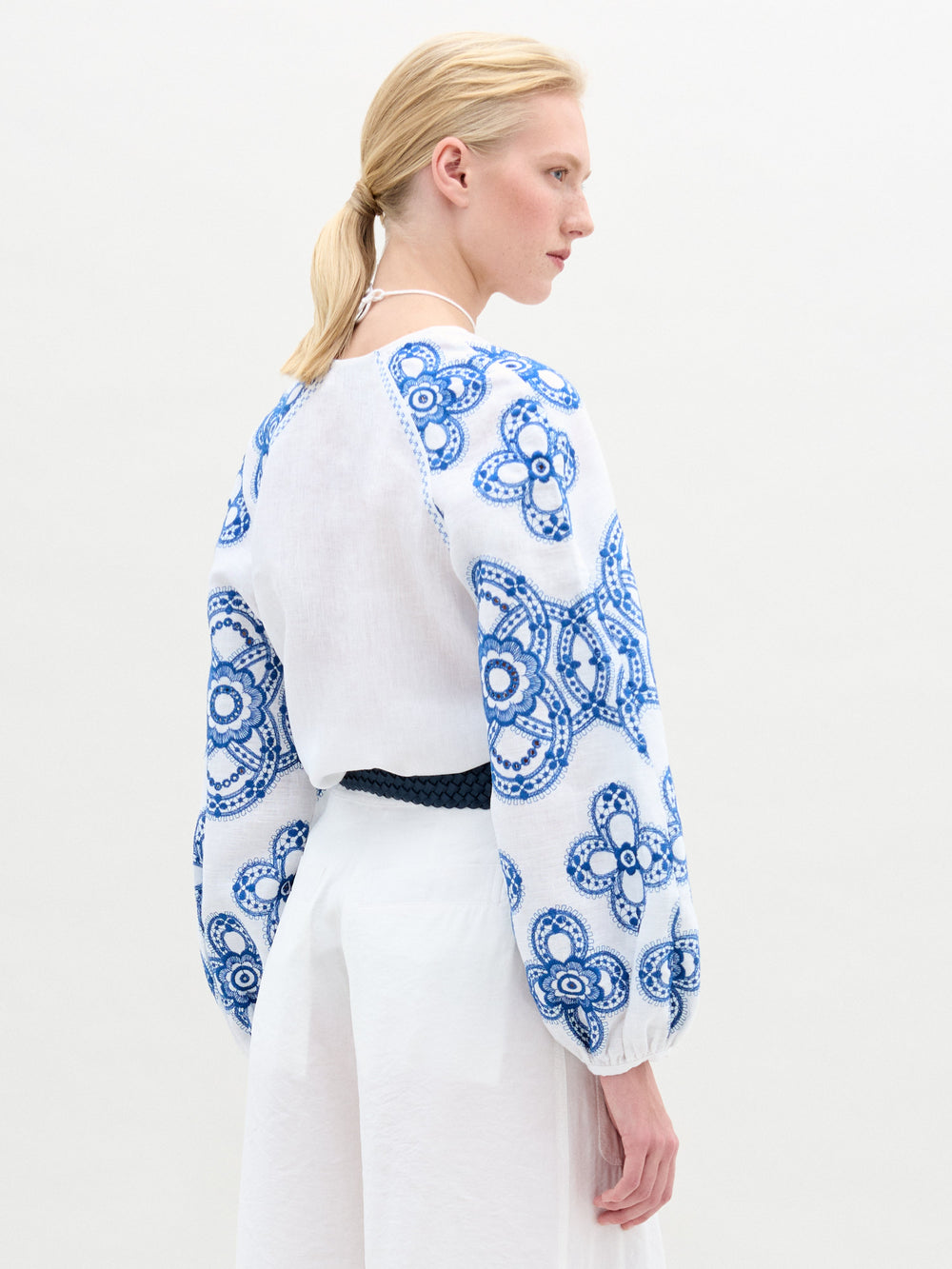 A woman stands against a plain background wearing the Molveno Blouse, an embroidered linen top with blue detailing and a V neckline, paired with a white skirt featuring a pocket and finished with a dark braided belt at her waist.